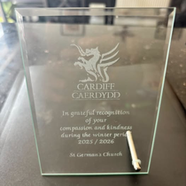 “A glass award plaque engraved with the Cardiff Council dragon emblem and bilingual text recognising St German’s Church for compassion and kindness during the 2025–2026 winter period, with a small metal pen resting against the base.