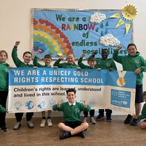 A group of primary‑school children in green uniforms stand and kneel in front of a colourful classroom display featuring a large paper rainbow, clouds and raindrops, and the words “We are a RAINBOW of endless possibilities.” They hold a wide UNICEF banner