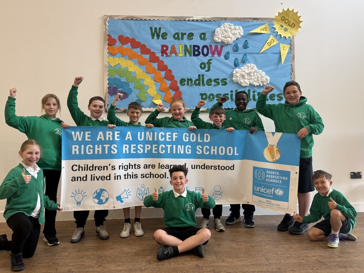 A group of primary‑school children in green uniforms stand and kneel in front of a colourful classroom display featuring a large paper rainbow, clouds and raindrops, and the words “We are a RAINBOW of endless possibilities.” They hold a wide UNICEF banner