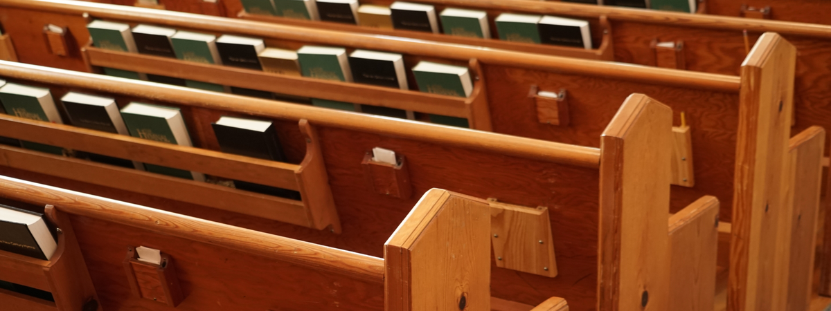 Empty wooden church pews, streamed in sunlight with green bibles on the shelves behind them