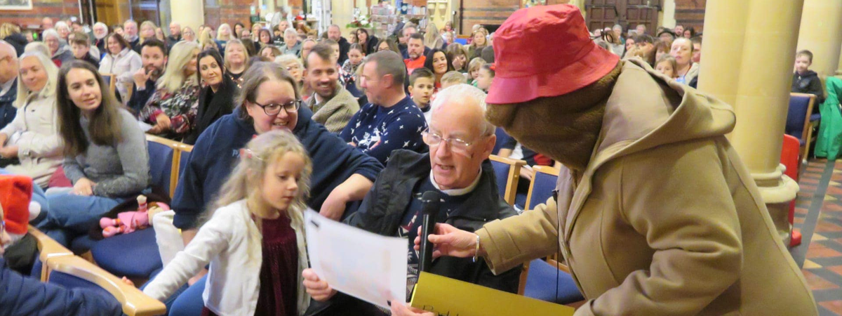 An adult dressed as Paddington Bear interviews an elderly gentleman and a small child.