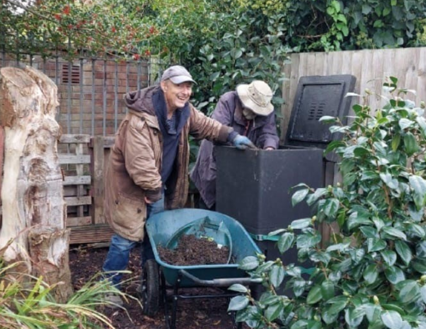 two male volunteers smiling while moving compost with a wheelbarrow in community garden